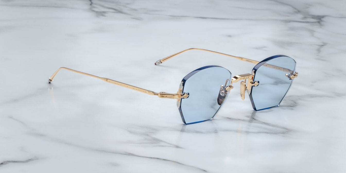 A pair of rimless, hexagonal gold eyeglasses with blue lenses and hand-painted 'tailfin' temple tips, displayed on a marble surface. side view