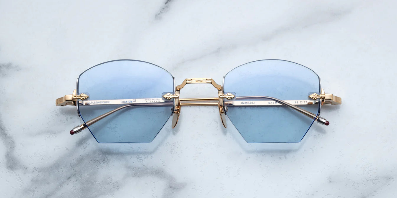A pair of rimless, hexagonal gold eyeglasses with blue lenses and hand-painted 'tailfin' temple tips, displayed on a marble surface.