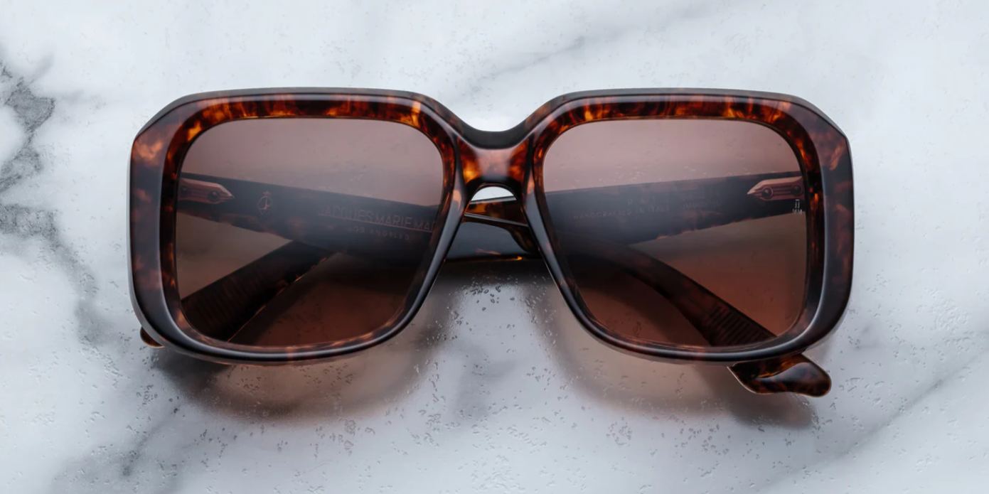 A pair of Jacques Marie Mage sunglasses with a refined, rectangular frame design, featuring a bow bridge and sculptural temples with polished metal details on a marble surface.