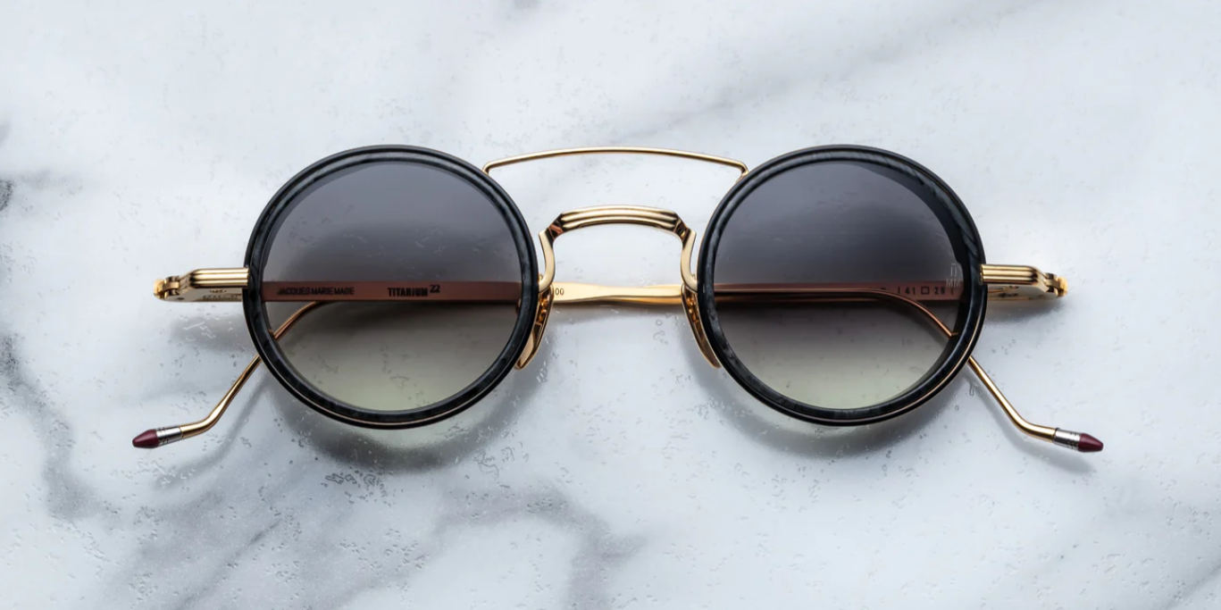 A pair of round full frame sunglasses with a double bridge design, acetate eye rim inserts, and hand-painted epoxy temple tips on a marble surface.