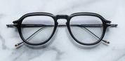A pair of black '50s-inspired aviator optical glasses with wirecore temples and a unique front-pin hinge design, laying on a marble surface.
