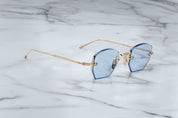 A pair of rimless, hexagonal gold eyeglasses with blue lenses and hand-painted 'tailfin' temple tips, displayed on a marble surface. side view
