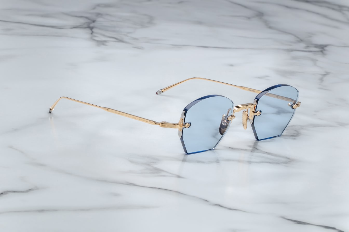 A pair of rimless, hexagonal gold eyeglasses with blue lenses and hand-painted 'tailfin' temple tips, displayed on a marble surface. side view