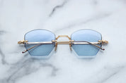 A pair of rimless, hexagonal gold eyeglasses with blue lenses and hand-painted 'tailfin' temple tips, displayed on a marble surface.