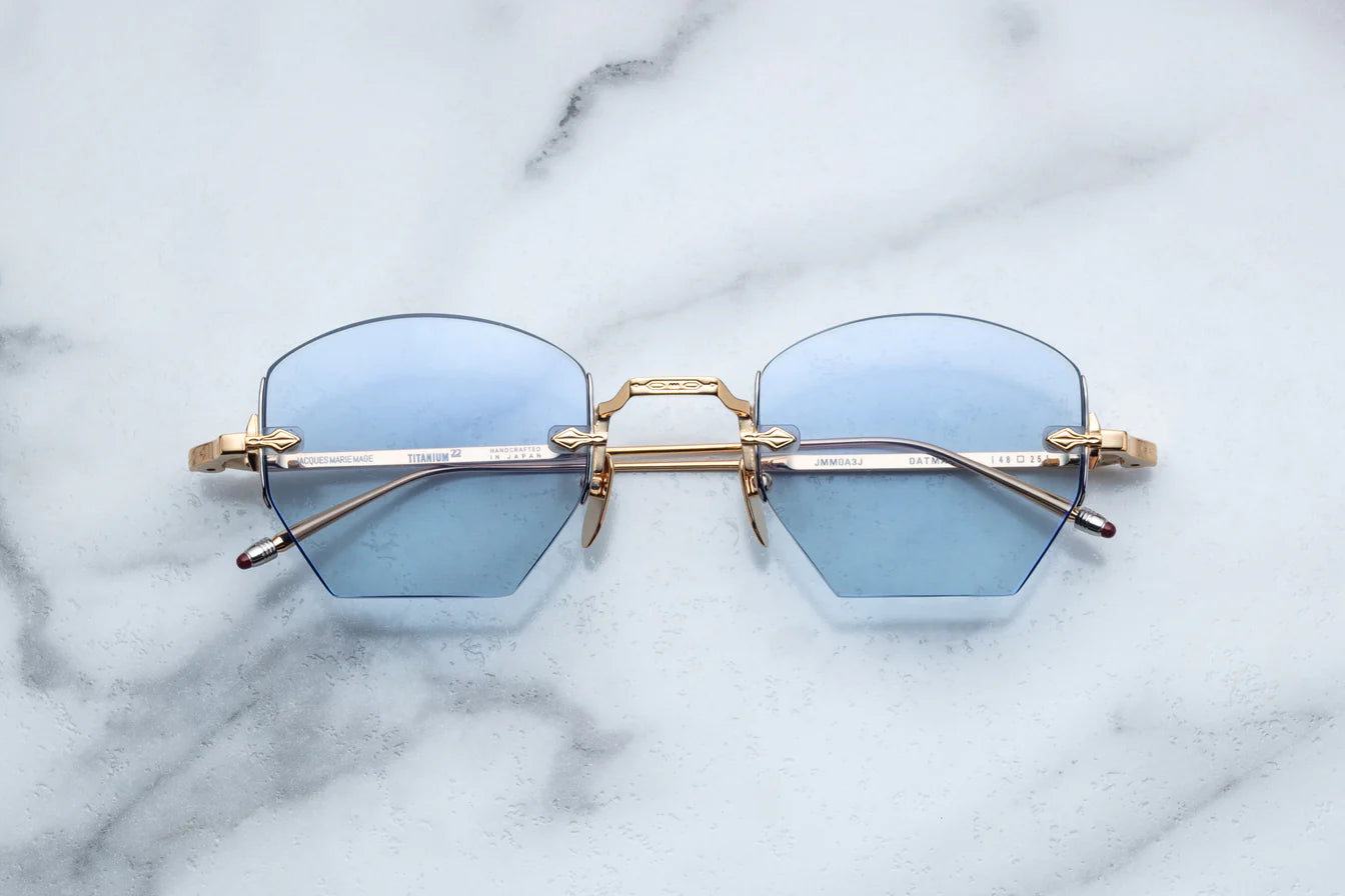 A pair of rimless, hexagonal gold eyeglasses with blue lenses and hand-painted 'tailfin' temple tips, displayed on a marble surface.