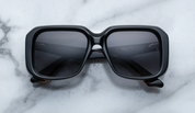 A pair of black, rectangular '70s-inspired sunglasses with sculptural temples and polished metal details, displayed on a marble surface.