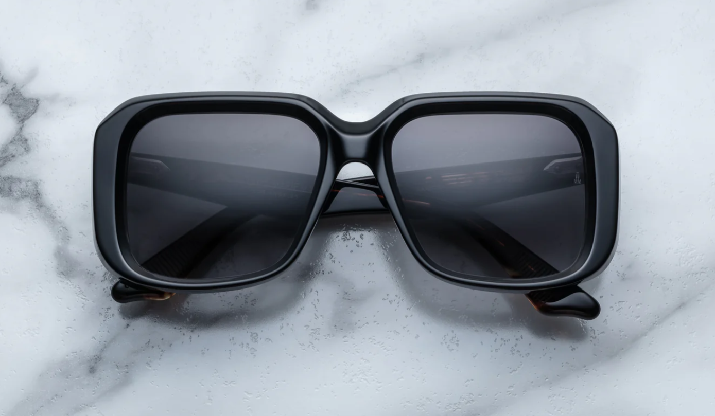 A pair of black, rectangular '70s-inspired sunglasses with sculptural temples and polished metal details, displayed on a marble surface.