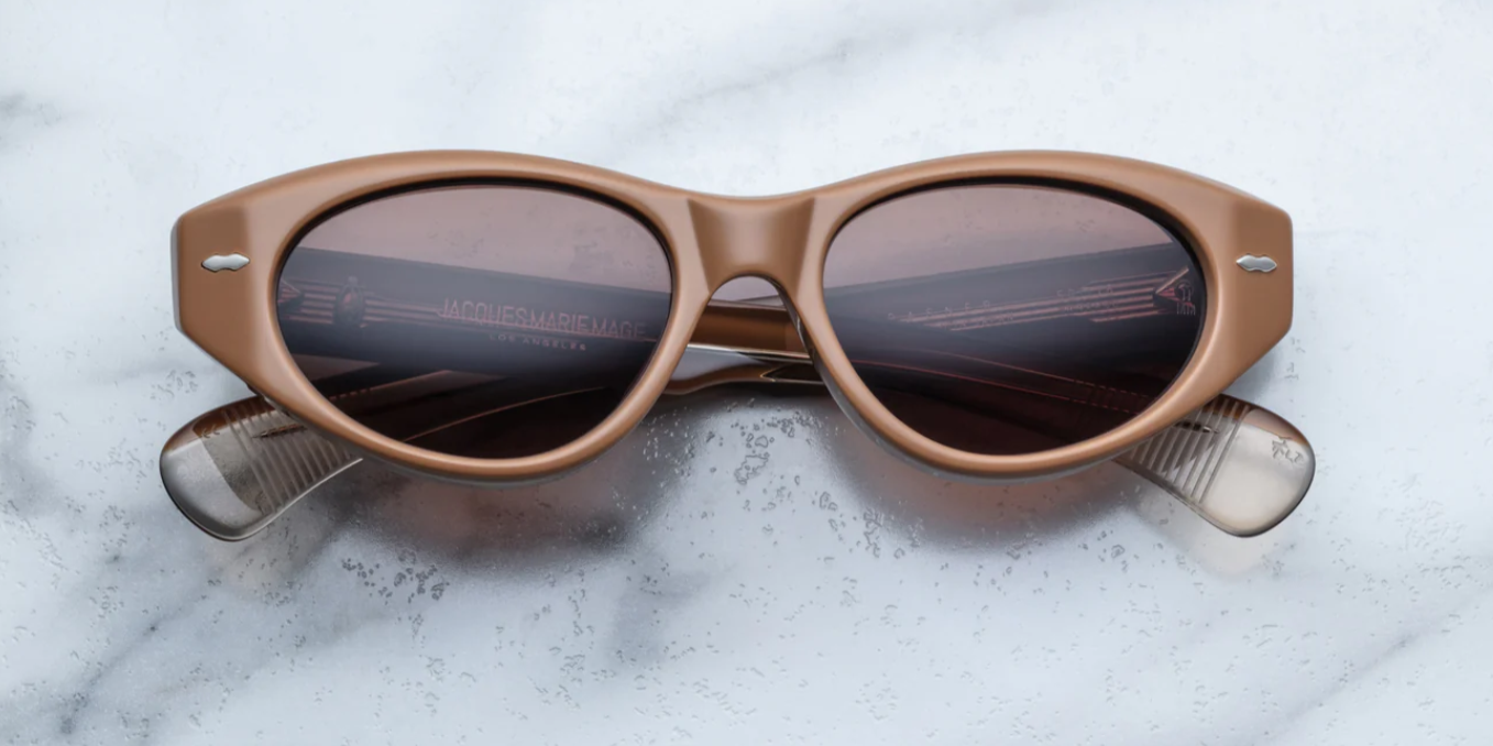 A pair of cat-eye style sunglasses with tobacco brown frame and lenses, featuring a double-arrowhead front pin and rounded paddle temples on a marble surface.