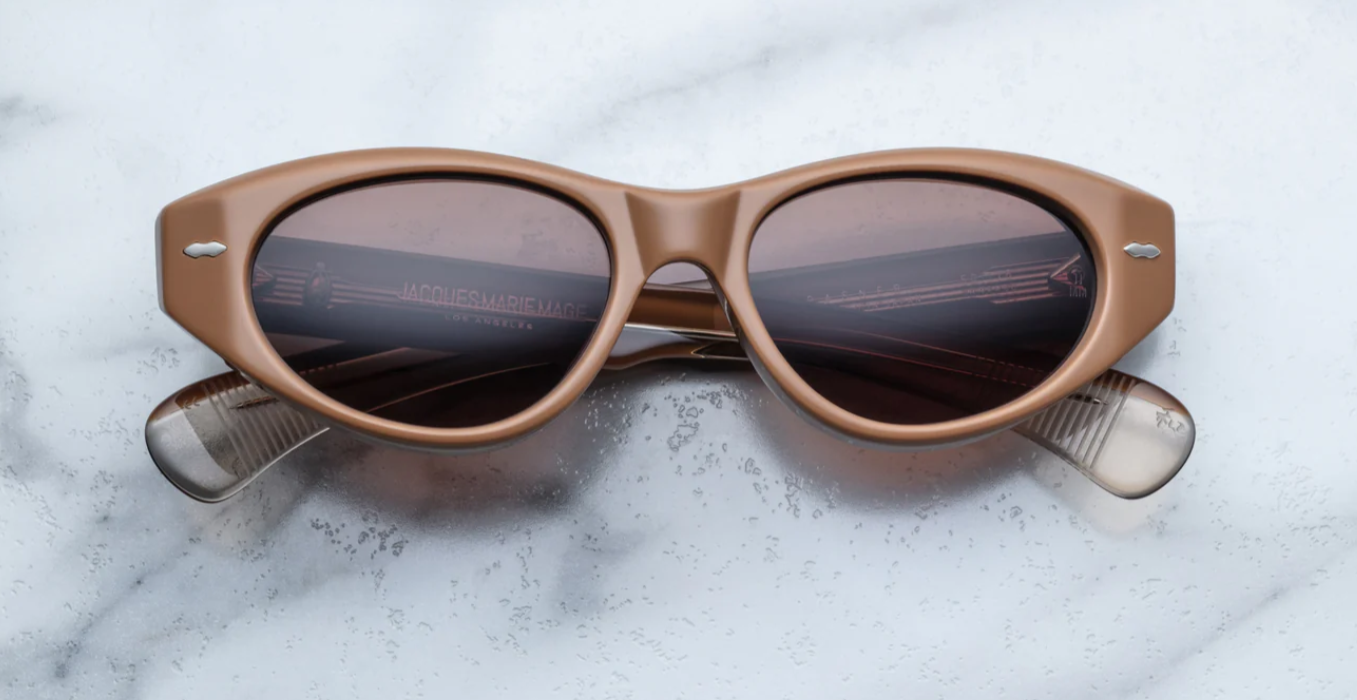 A pair of cat-eye style sunglasses with tobacco brown frame and lenses, featuring a double-arrowhead front pin and rounded paddle temples on a marble surface.
