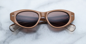 A pair of cat-eye style sunglasses with tobacco brown frame and lenses, featuring a double-arrowhead front pin and rounded paddle temples on a marble surface.