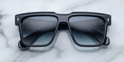 A pair of black rectangular framed sunglasses with a sculptural brow, resting on a marble surface.