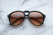 A pair of '80s-inspired oversized aviator sunglasses with a black frame and bronze-colored lenses, placed on a marble surface.