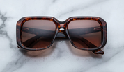 A pair of Jacques Marie Mage sunglasses with a refined, rectangular frame design, featuring a bow bridge and sculptural temples with polished metal details on a marble surface.
