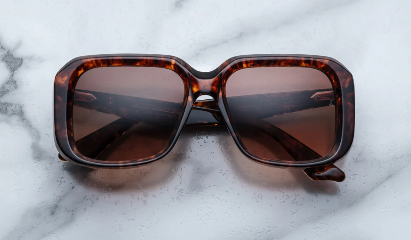 A pair of Jacques Marie Mage sunglasses with a refined, rectangular frame design, featuring a bow bridge and sculptural temples with polished metal details on a marble surface.