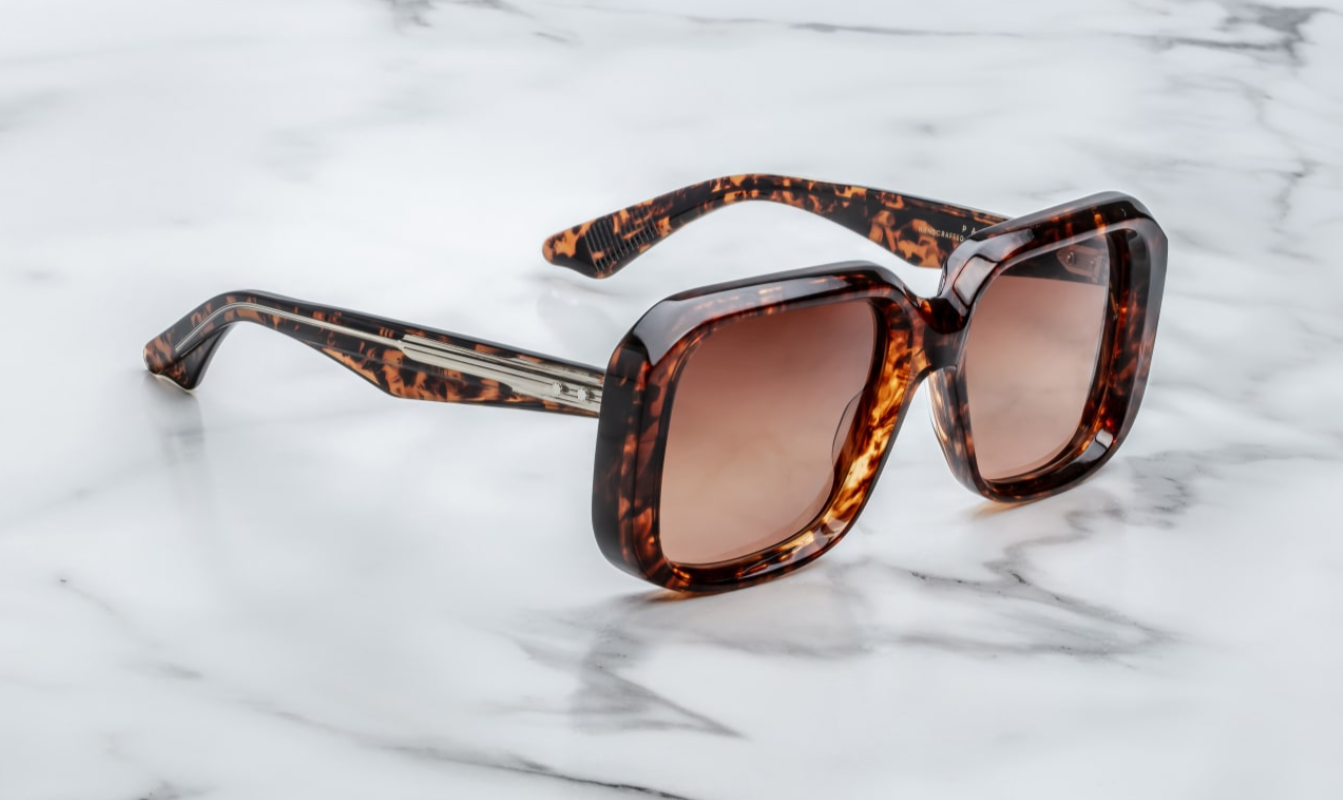 A pair of Jacques Marie Mage sunglasses with a refined, rectangular frame design, featuring a bow bridge and sculptural temples with polished metal details on a marble surface. side view