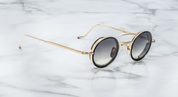 A pair of round full frame sunglasses with a double bridge design, acetate eye rim inserts, and hand-painted epoxy temple tips on a marble surface.