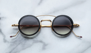 A pair of round full frame sunglasses with a double bridge design, acetate eye rim inserts, and hand-painted epoxy temple tips on a marble surface.