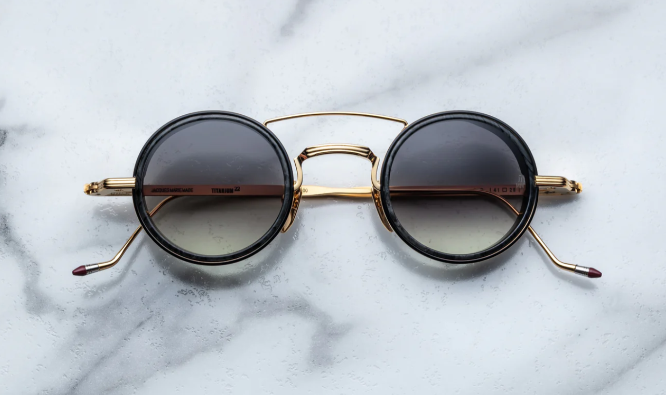 A pair of round full frame sunglasses with a double bridge design, acetate eye rim inserts, and hand-painted epoxy temple tips on a marble surface.