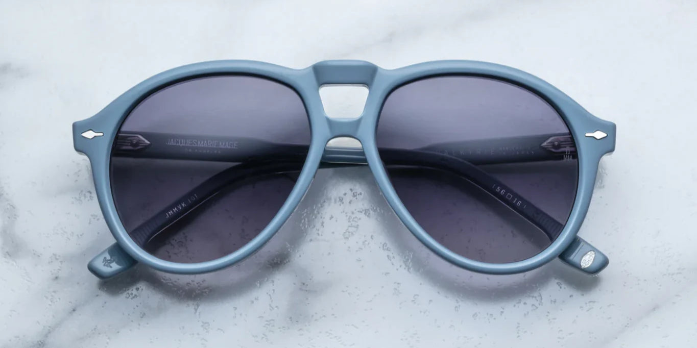 A pair of '80s-inspired oversized aviator sunglasses with a blue frame and blue-colored lenses, placed on a marble surface.