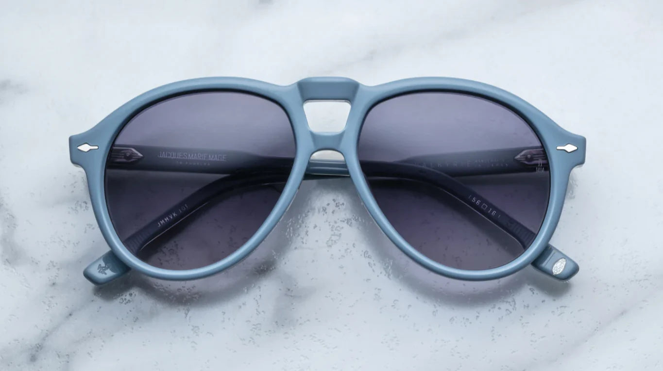 A pair of '80s-inspired oversized aviator sunglasses with a blue frame and blue-colored lenses, placed on a marble surface.