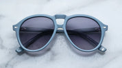 A pair of '80s-inspired oversized aviator sunglasses with a blue frame and blue-colored lenses, placed on a marble surface.