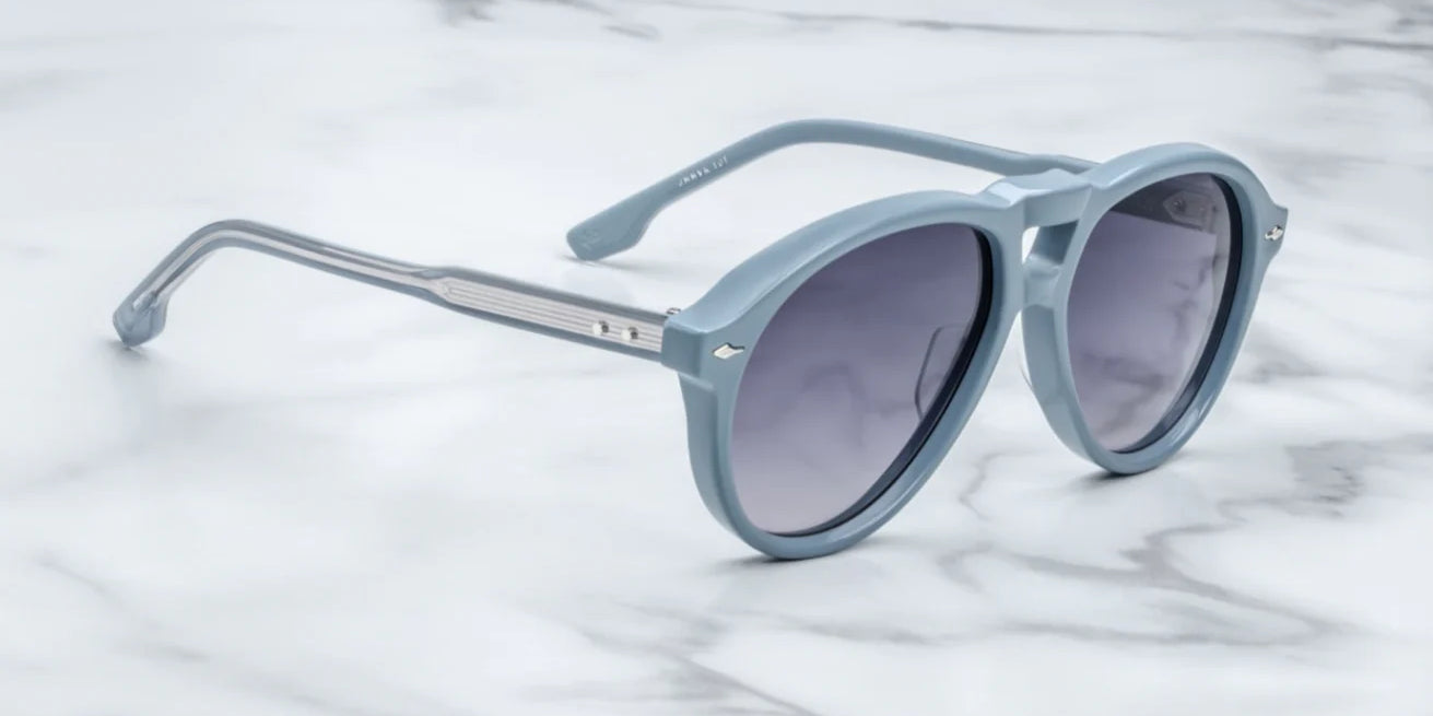 A pair of '80s-inspired oversized aviator sunglasses with a blue frame and blue-colored lenses, placed on a marble surface. side view