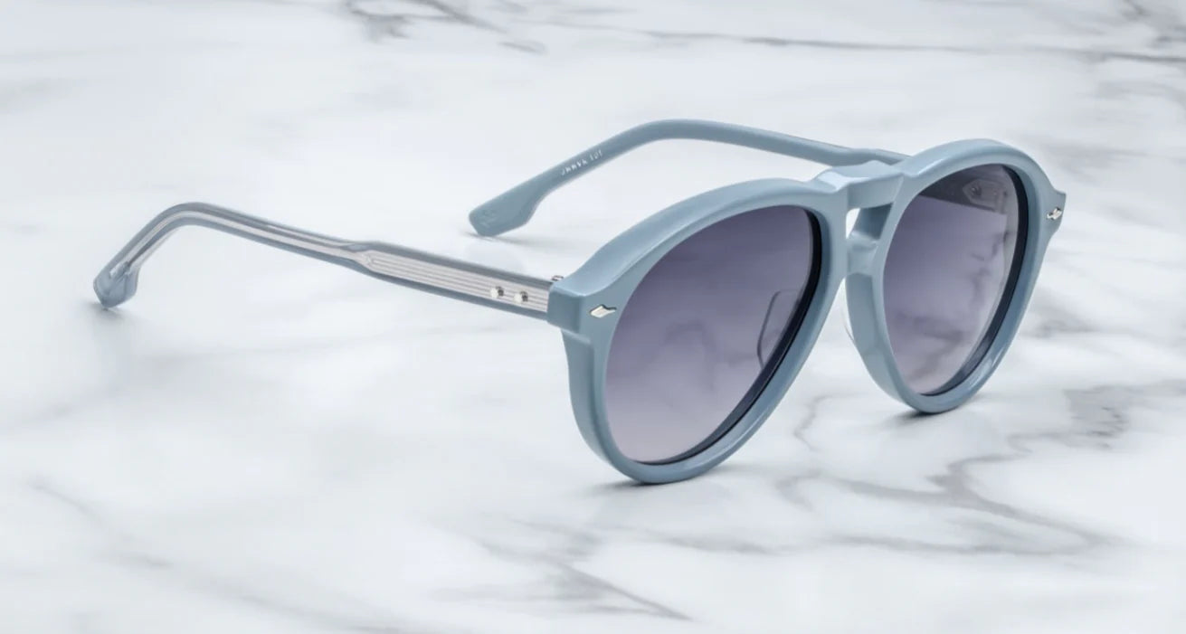 A pair of '80s-inspired oversized aviator sunglasses with a blue frame and blue-colored lenses, placed on a marble surface. side view